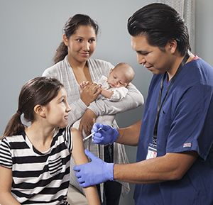 Health care provider giving girl injection in arm while woman with baby looks on.
