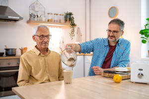 At their kitchen table, a caretaker helps pour a drink for an older family member.