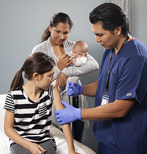 Health care provider giving girl injection in arm while woman holding baby looks on.